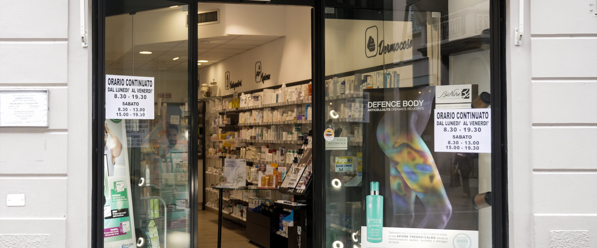Ingresso della Farmacia Corvi di Piacenza con le vetrine e l’orario di apertura